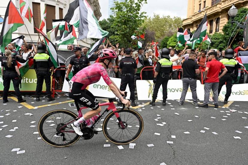 A cyclist competes as pro-Palestinian protesters holding Palestinian and Basque flags demonstrate during the eleventh stage of the Vuelta a Espana cycling tour, a 167 km race from Bilbao to Bilbao, on September 3, 2025.  ANDER GILLENEA / AFP